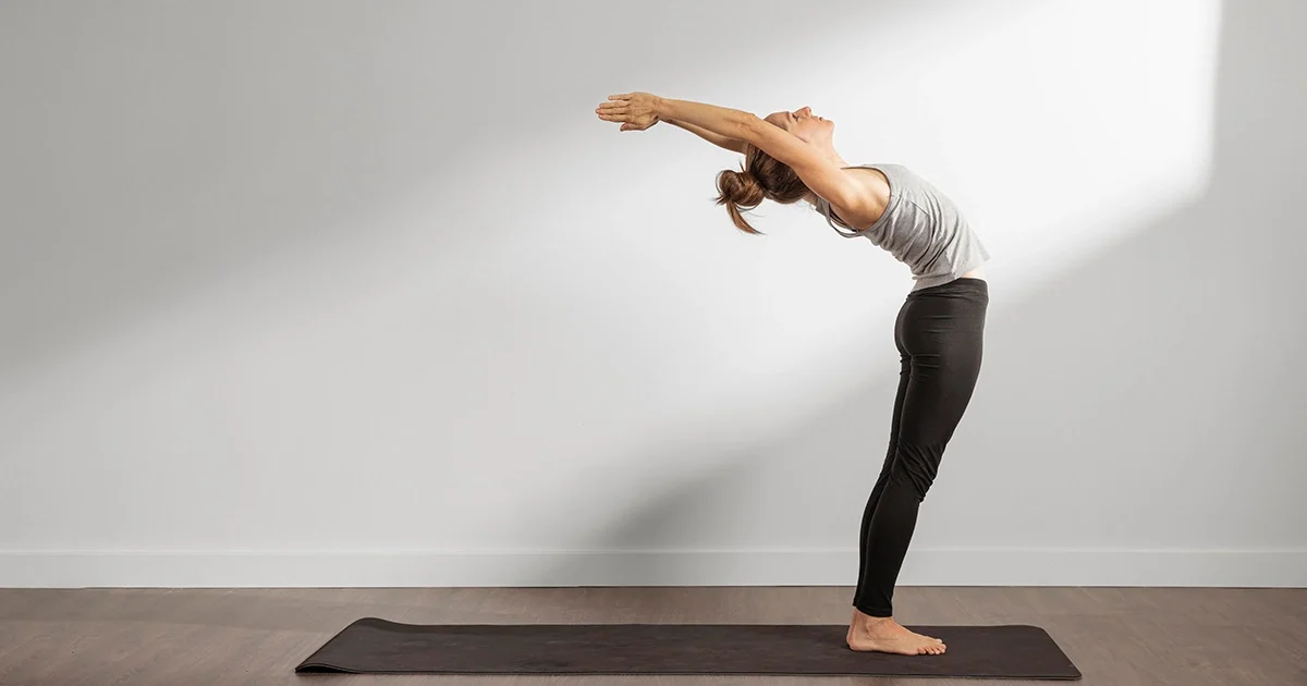 A practitioner demonstrates Ardha Chakrasana by supporting the lower back with both hands while gently arching the upper spine backward.