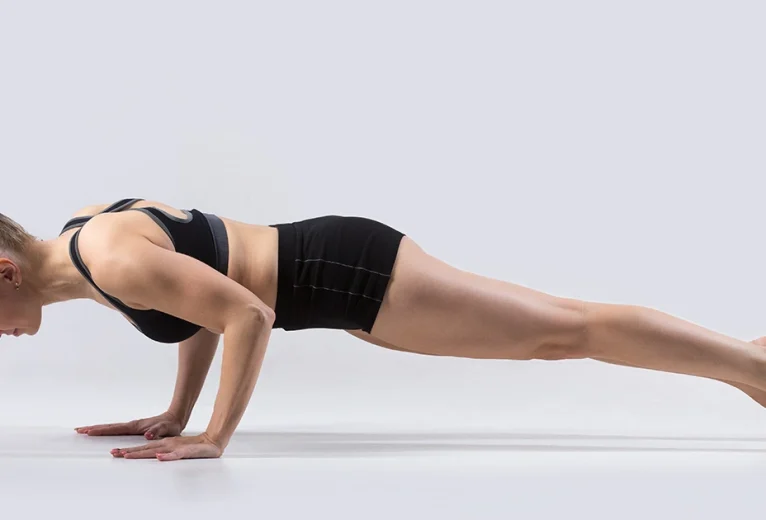 A practitioner demonstrates the correct hover alignment of Chaturanga Dandasana on a blue mat, maintaining the shoulders at elbow height.