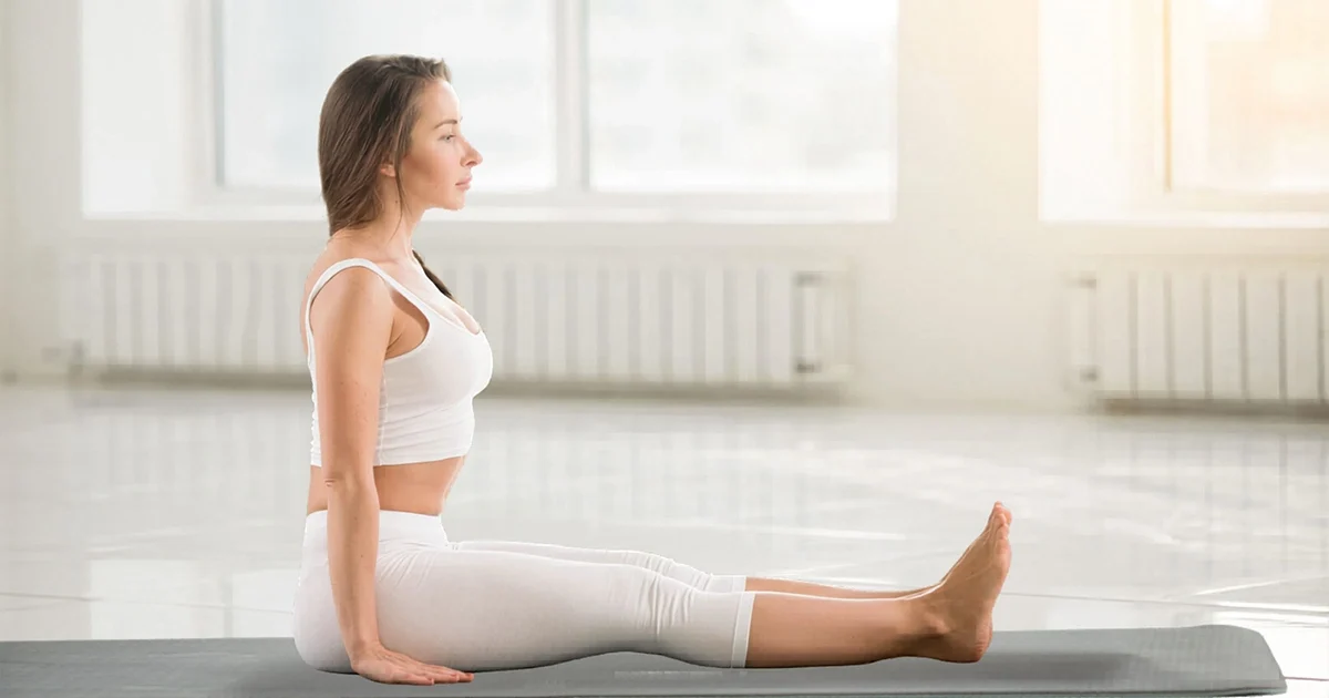 A practitioner demonstrates the correct alignment of Dandasana on a blue mat, showcasing a long vertical spine and fully extended legs.