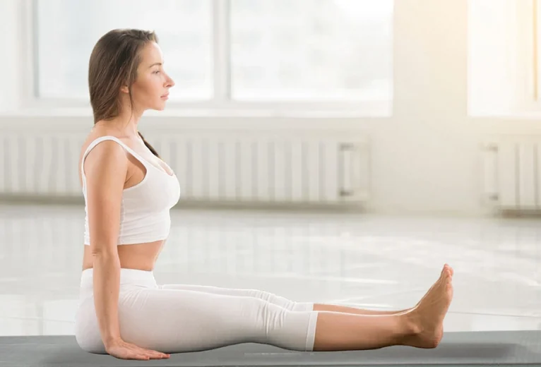 A practitioner demonstrates the correct alignment of Dandasana on a blue mat, showcasing a long vertical spine and fully extended legs.