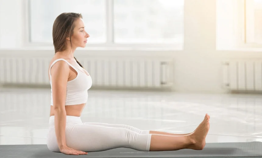 A practitioner demonstrates the correct alignment of Dandasana on a blue mat, showcasing a long vertical spine and fully extended legs.