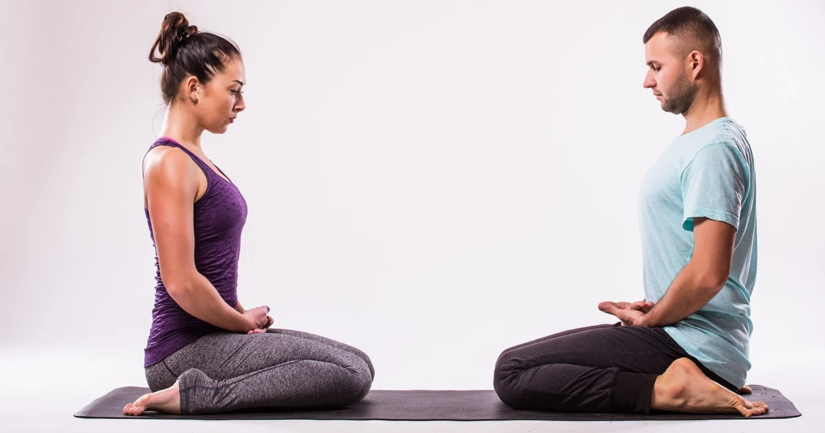 A practitioner demonstrates proper alignment in Virasana on a purple mat, using a yoga block under the hips to make Hero Pose more comfortable.