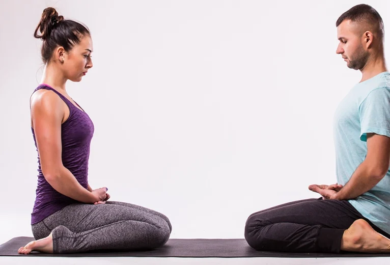 A practitioner demonstrates proper alignment in Virasana on a purple mat, using a yoga block under the hips to make Hero Pose more comfortable.