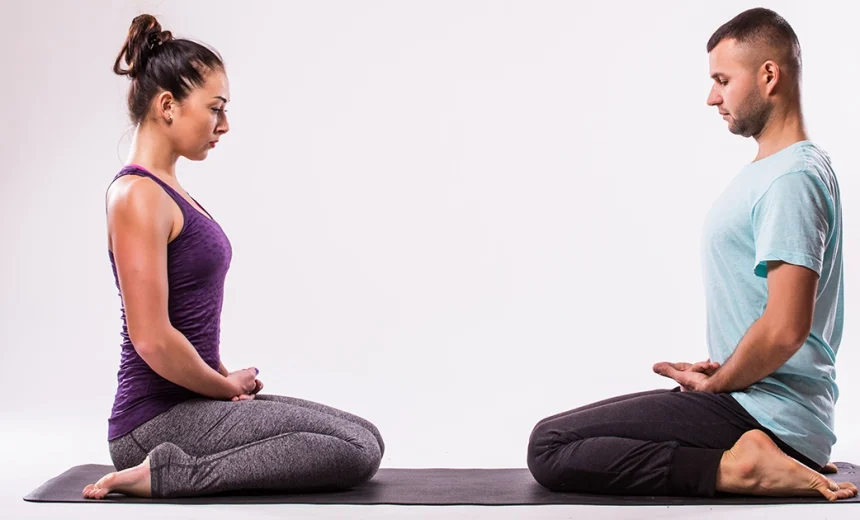 A practitioner demonstrates proper alignment in Virasana on a purple mat, using a yoga block under the hips to make Hero Pose more comfortable.